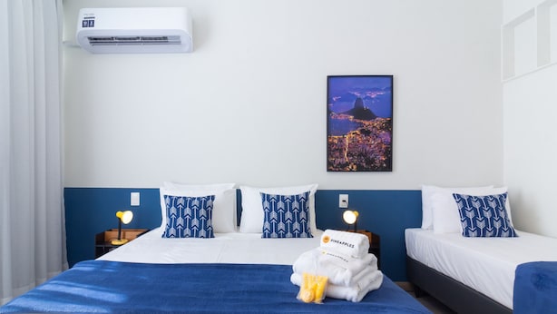Ein Hotelzimmer mit einem großen Bett, zwei Nachttischen, einer fest installierten Klimaanlage und einem Stadtbild in einem Bilderrahmen.