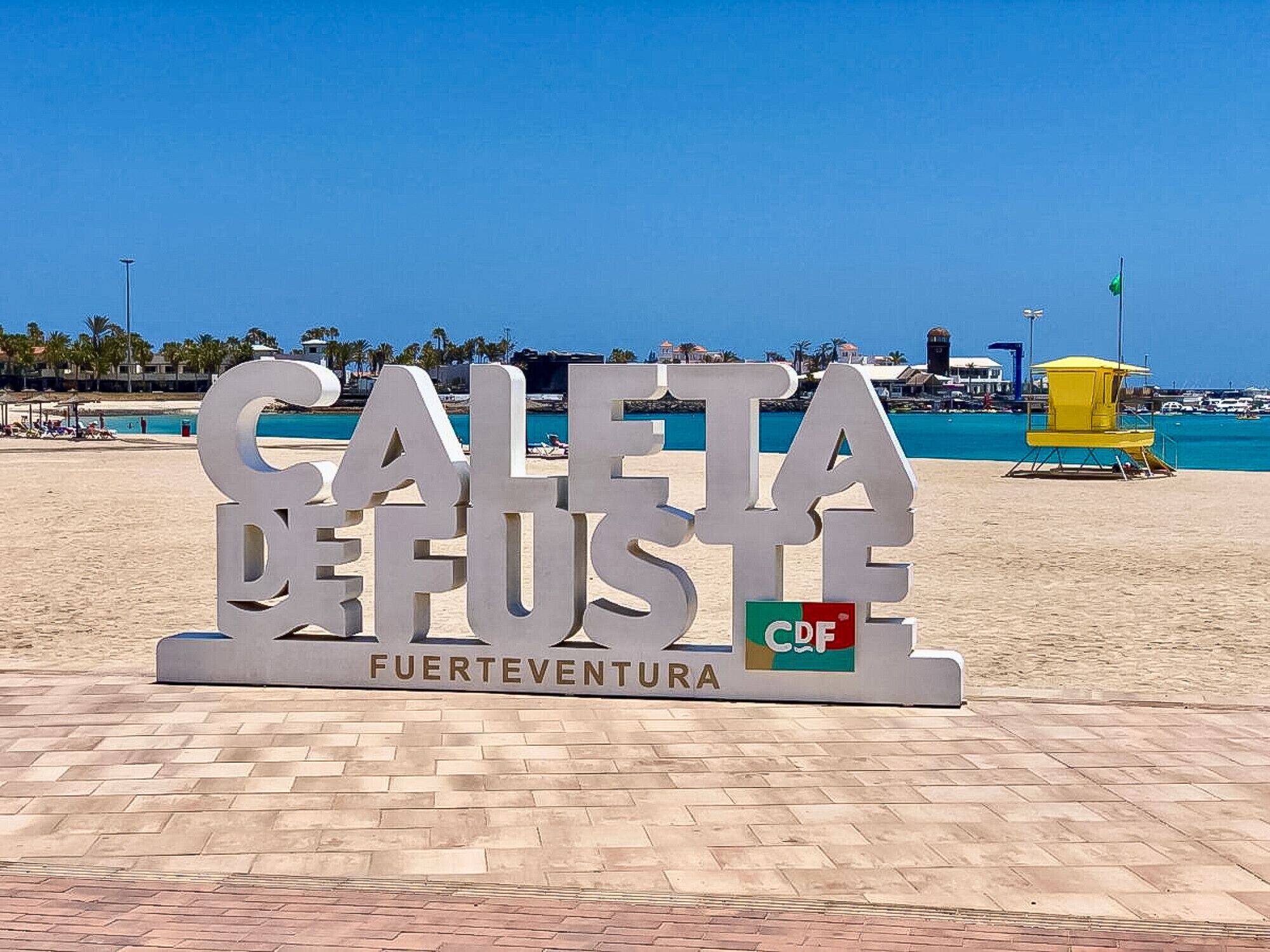 Un cartel de playa con el texto "CAETA DE FUSTE" y "FUERTEVENTURA" indica un lugar en una playa soleada con cielos despejados y una torre de salvavidas al fondo.