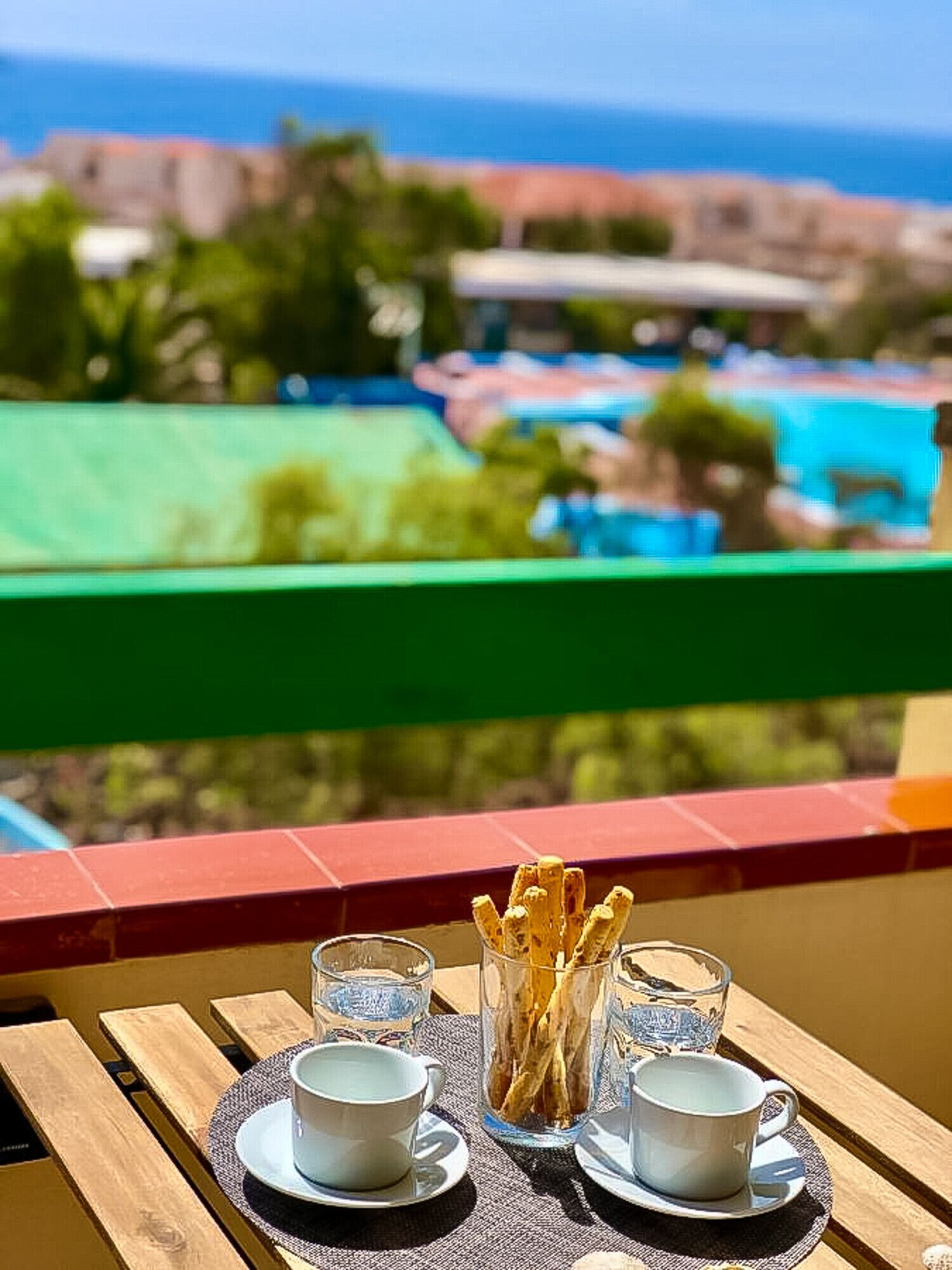 A table with two cups and a glass of water, and a glass with sticks, set against a view of the sea and buildings.