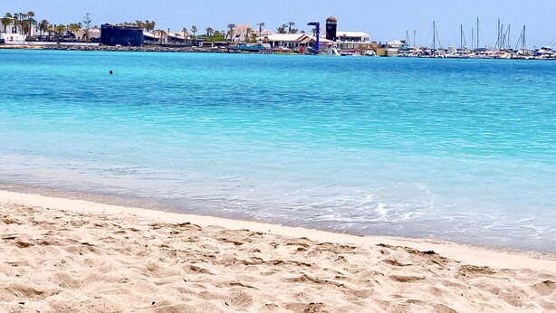 Una playa con agua turquesa cristalina, una orilla arenosa y un puerto deportivo con barcos.