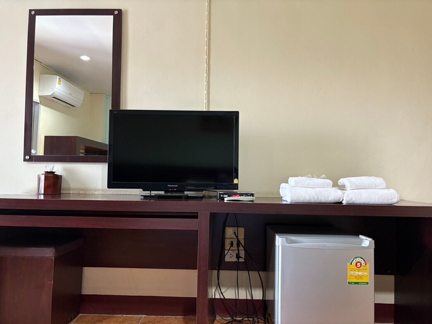 Habitación de hotel con televisor de pantalla plana, refrigerador pequeño, espejo y toallas dobladas ordenadamente sobre un escritorio de madera.