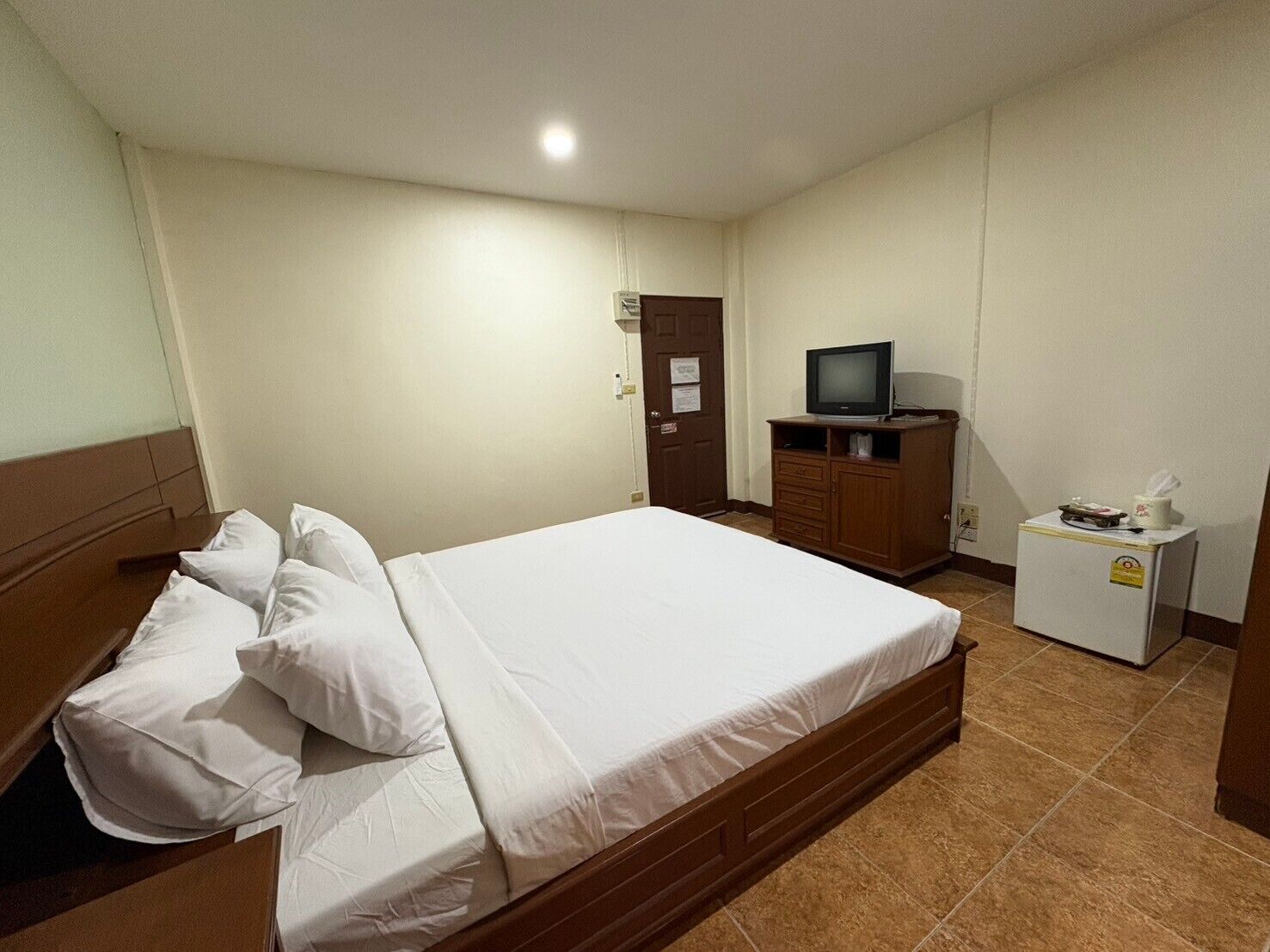 A hotel room with a single bed, a wooden headboard, a small wooden cabinet with a television, and a mini-fridge.