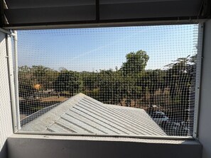 A view through a window with a metal mesh, overlooking a rooftop with a corrugated metal roof and trees in the background.