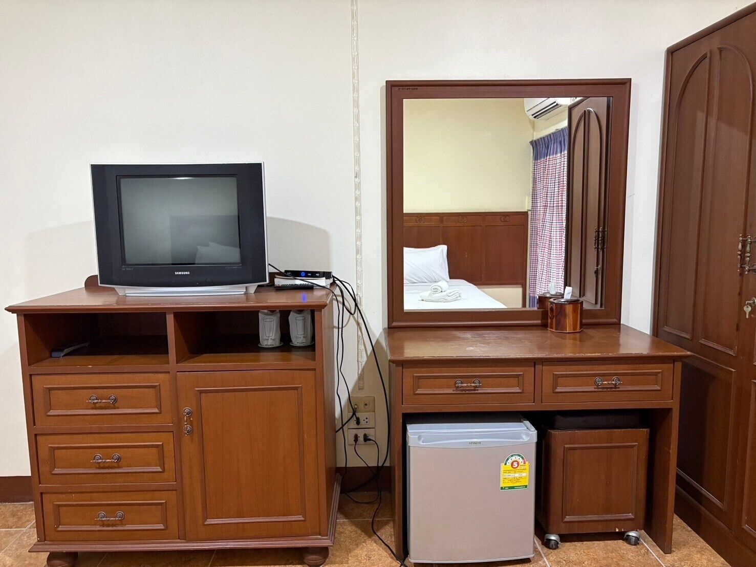 A hotel room with a wooden desk, a television, a mini-fridge, and a large mirror reflecting the room.