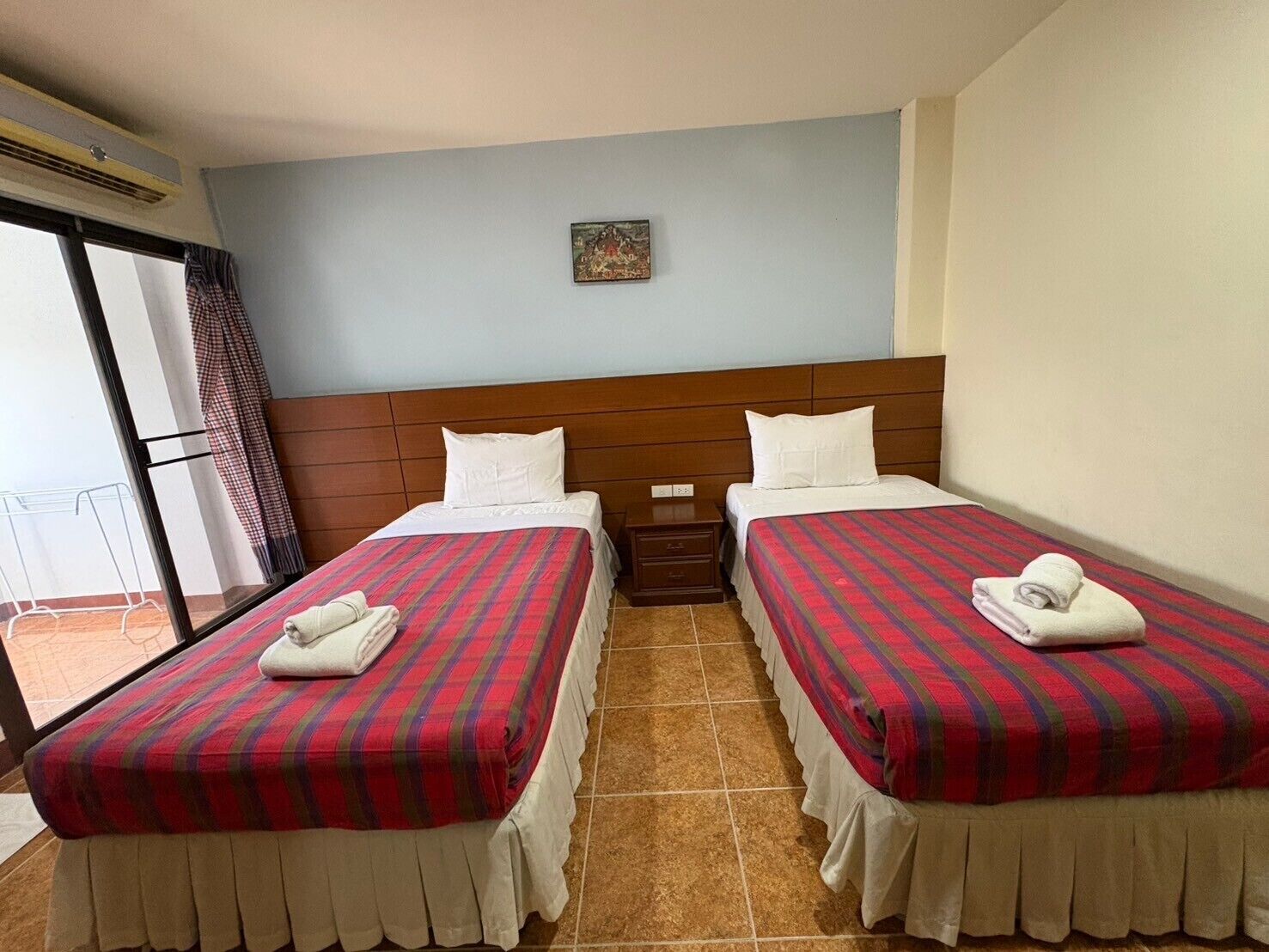 Habitación de hotel con dos camas, cada una con colcha a cuadros blancos y rojos, almohadas blancas y una toalla blanca doblada. Hay una mesita de noche pequeña entre las camas y una ventana con cortina a la izquierda.