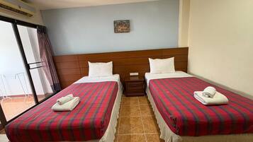 Habitación de hotel con dos camas, cada una con colcha a cuadros blancos y rojos, almohadas blancas y una toalla blanca doblada. Hay una mesita de noche pequeña entre las camas y una ventana con cortina a la izquierda.