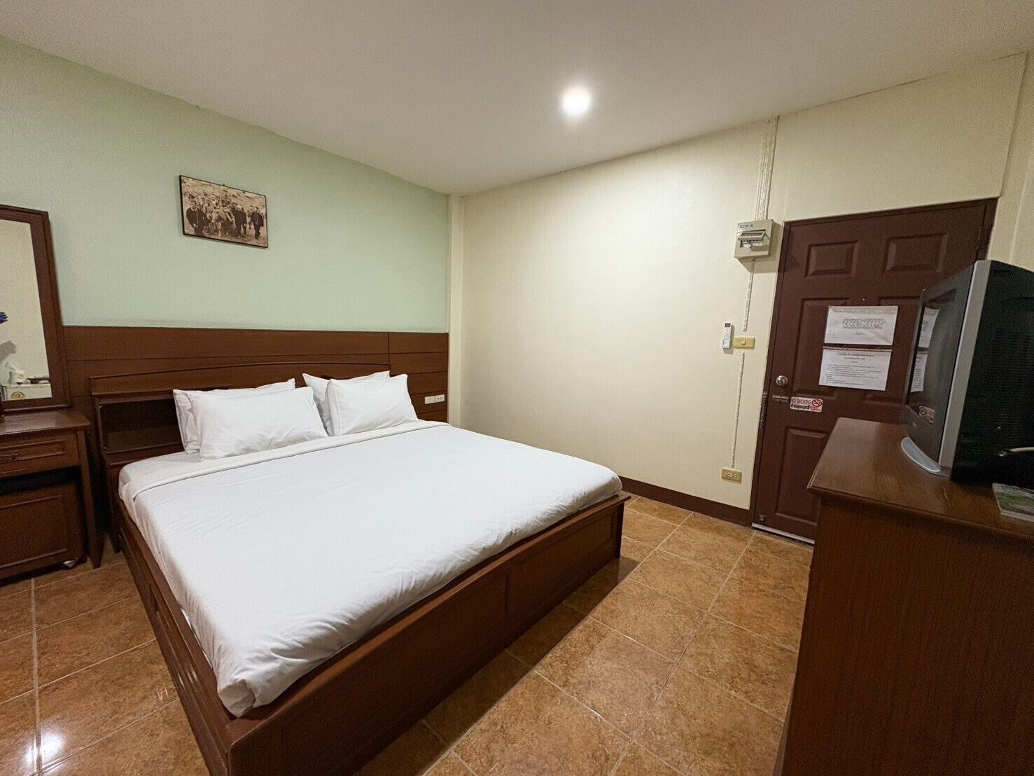 A hotel room with a single bed, a wooden nightstand, a desk with a computer, a television, and a door with a sign.
