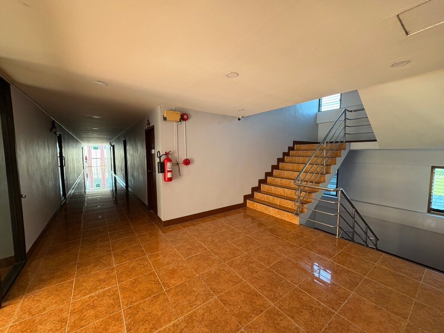 Um corredor amplo com um extintor fixado na parede, uma escada com grades de metal e piso em azulejo.
