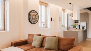 A modern living room with a brown sofa, a small table with a remote and a phone, a decorative wall clock, and a kitchen in the background. - Casa da Eira - Rural Retreat (Vilar)