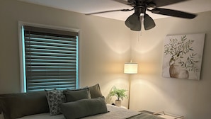 A bedroom with a bed, a ceiling fan, a bedside lamp, a window with blinds, and a wall-mounted plant.