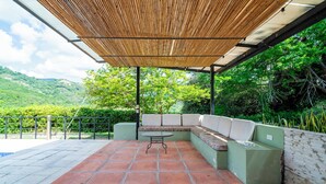 Een terras buiten met een bamboe overkapping, een ronde tafel en een ingebouwde bank met kussens.