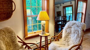 A room with two wicker chairs, a lamp, a window with a view of greenery, and a mirror reflecting the room. - Historic Edge of the River, Adirondacks (Willsboro)