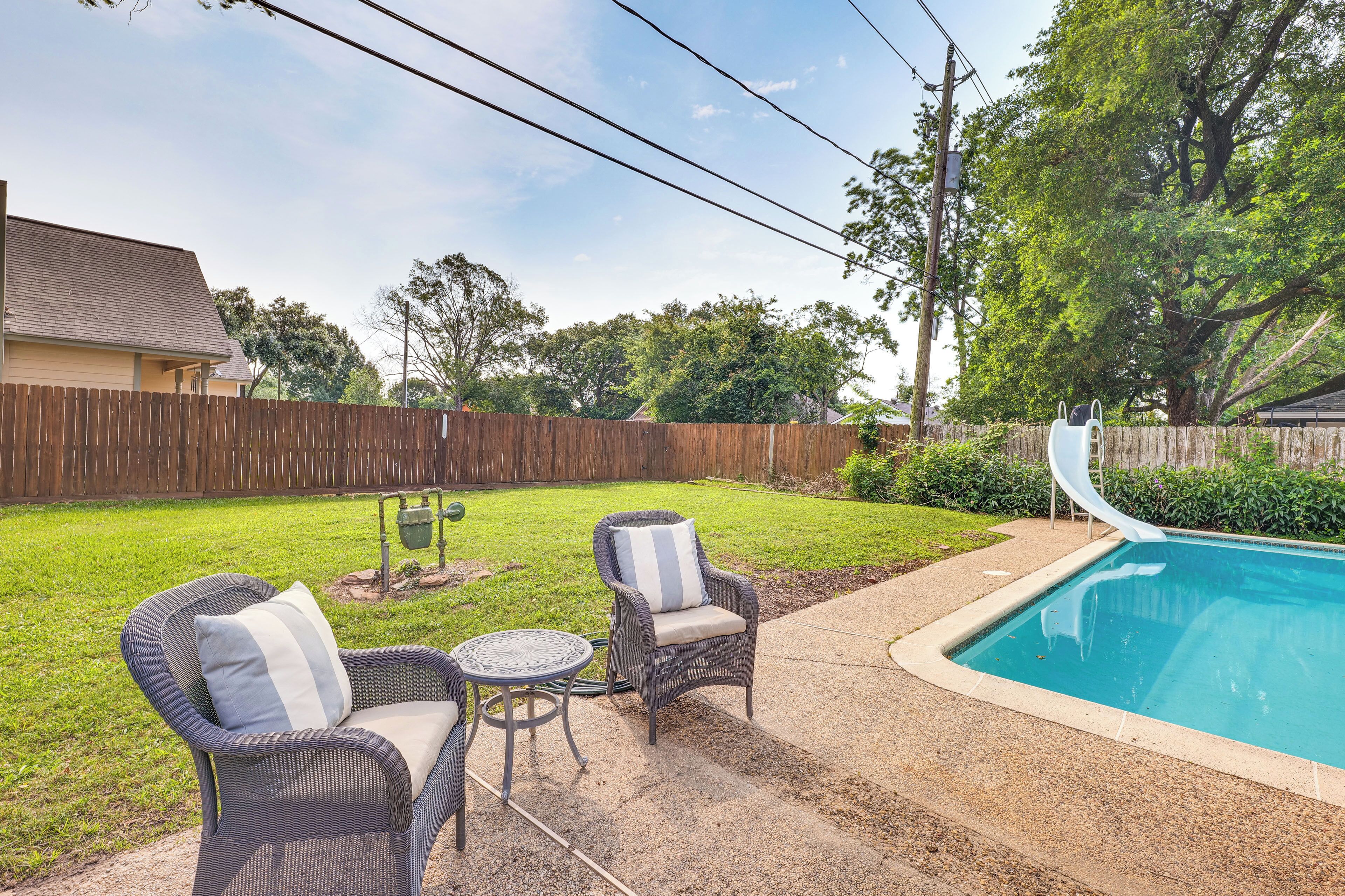 Un jardin avec une piscine, deux fauteuils en osier dotés de coussins, une petite table ronde et un toboggan.