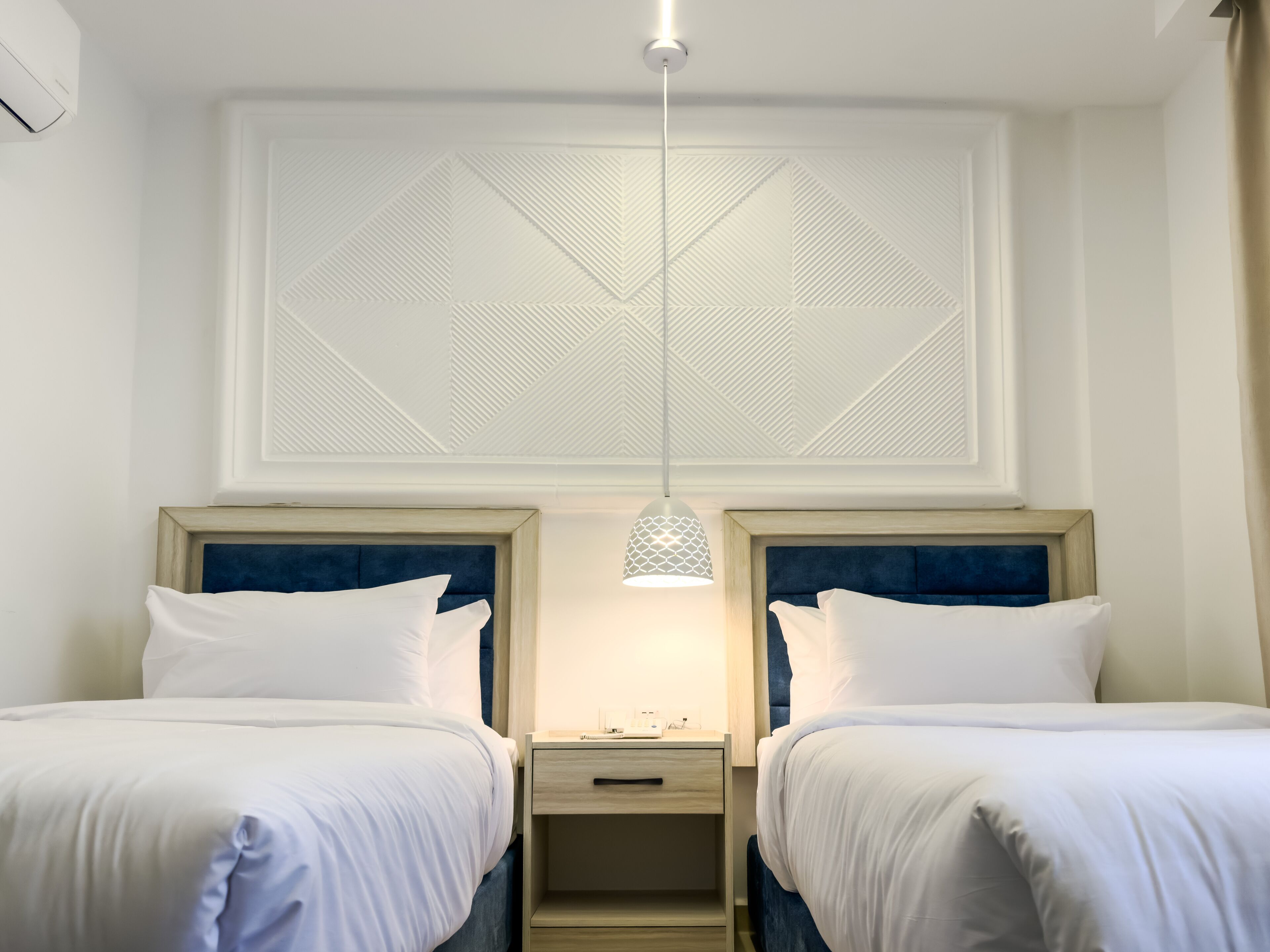 A hotel room with two beds, a nightstand, a pendant light, and a geometric-patterned ceiling.