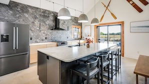 A modern kitchen with a large island, stainless steel appliances, and a stone backsplash. There are two hanging wooden paddles on the wall and a window with a view of the outdoors. - Modern Cabin Near Cheney Lake W Pool and Hot Tub - Holiday Home (Pretty Prairie)