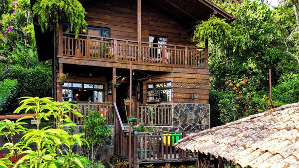 A wooden two-story house with a balcony, surrounded by lush greenery and a stone pathway leading to the entrance. - Charming 2-bedroom cabin in private condominium (Juayua)