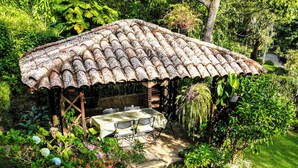 A rustic outdoor patio with a tiled roof, surrounded by lush greenery and various plants. - Charming 2-bedroom cabin in private condominium (Juayua)