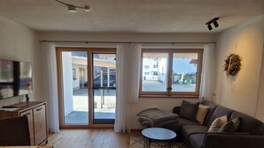 A living room with a sofa, a small round table, a TV on a wooden cabinet, and a sliding glass door leading to an outdoor area.