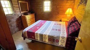 A wooden cabin bedroom with a bed, dresser, two windows, and a lamp. - Cold Spring Lodge - Cabin 5 (Big Indian)