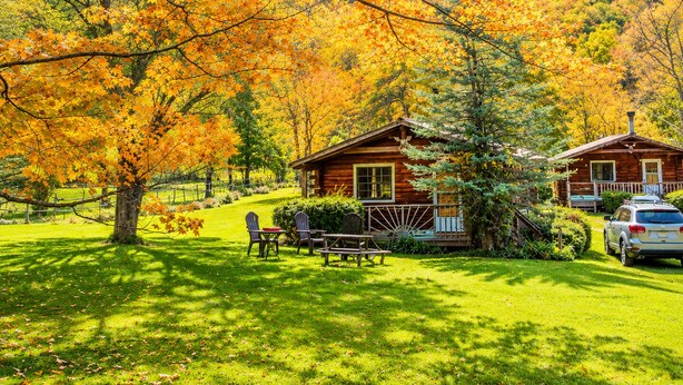 A cabin with a deck, surrounded by a grassy area and trees with autumn leaves. - Cold Spring Lodge - Cabin 5 (Big Indian)