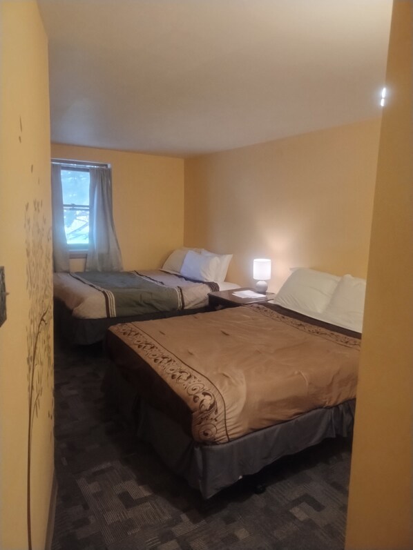 A hotel room with two beds, a window with curtains, a bedside table with a lamp, and a patterned carpet.