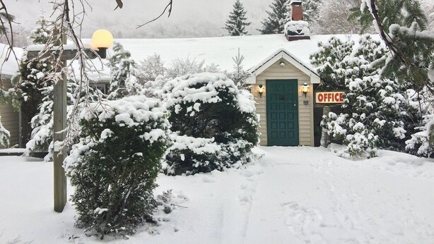 Un piccolo edificio coperto di neve, con una porta verde e un cartello con la scritta 'OFFICE', circondato da alberi carichi di neve e un cielo nuvoloso.