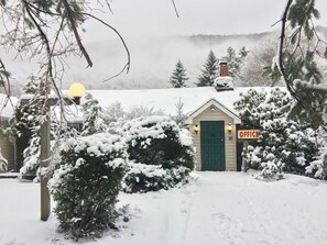 A small, snow-covered building with a green door and an 'OFFICE' sign, surrounded by snow-laden trees and a cloudy sky.