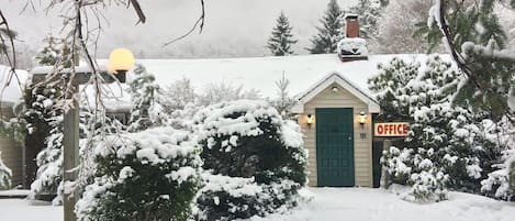 A small, snow-covered building with a green door and an 'OFFICE' sign, surrounded by snow-laden trees and a cloudy sky.