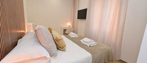 A hotel room with a bed, a bedside table, a TV mounted on the wall, and a window with curtains.