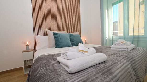 Una cama bien hecha con toallas dobladas, una mesita de noche con una lámpara y un cabecero de madera.