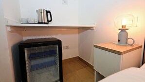 Habitación de hotel compacta con refrigerador, un mueble pequeño, una repisa con tetera y tazas, y una mesita de noche con lámpara.