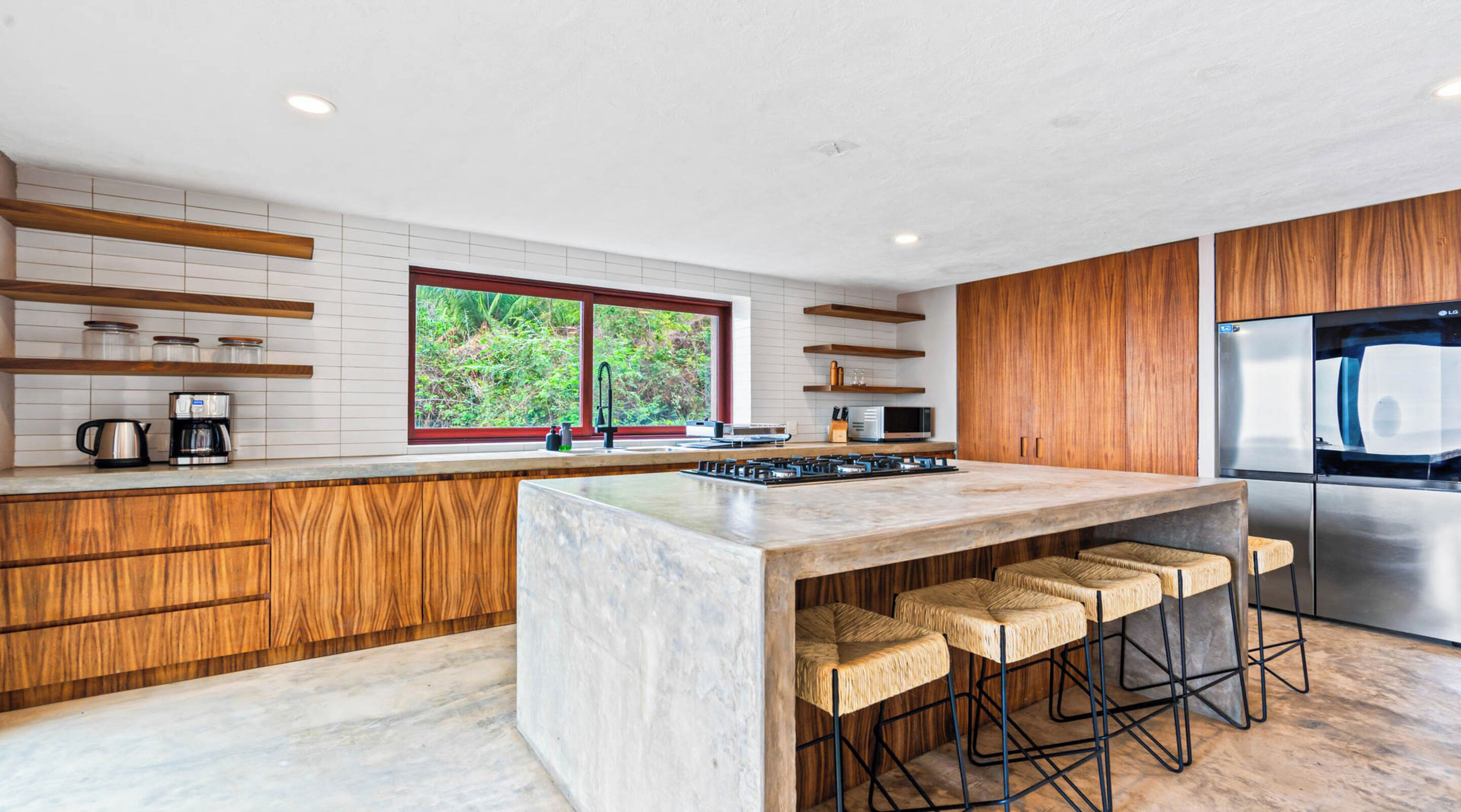 A modern kitchen with a central island, wooden cabinetry, a stainless steel refrigerator, and a window with a view of greenery.
