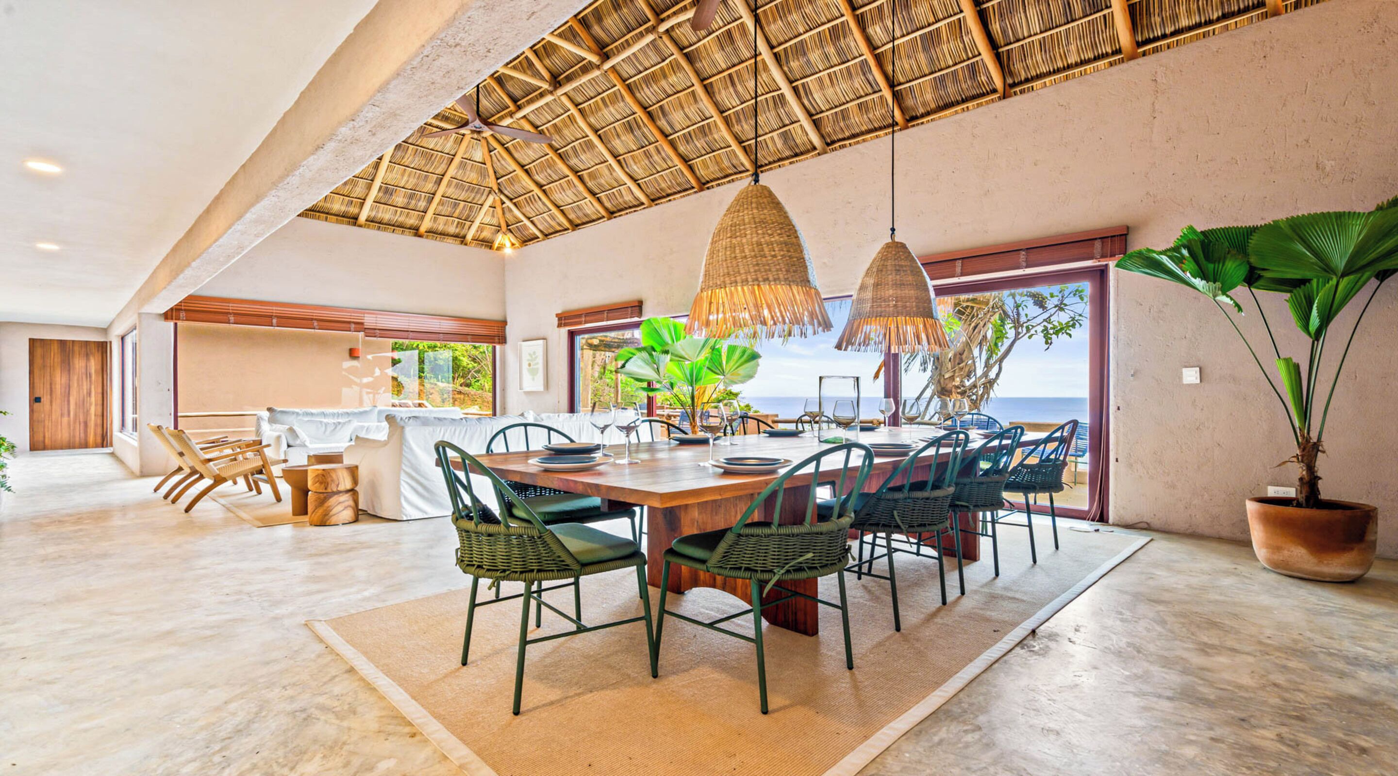 A spacious dining area with a large wooden table, green chairs, and a thatched ceiling.