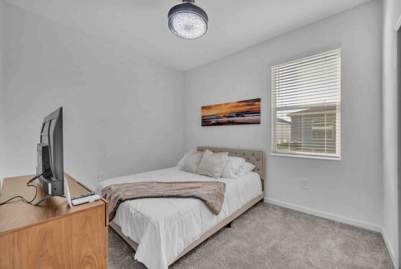 Une chambre à coucher avec un lit, un bureau avec un ordinateur, un tableau accroché au mur et une fenêtre avec des stores.