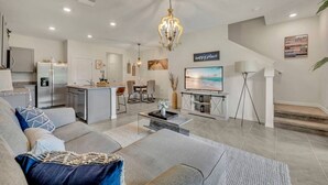 Una sala de estar moderna con un sofá, una mesa de centro y un televisor. Se observa una zona de cocina con un refrigerador y una mesa de comedor. En el fondo, se ve una escalera.