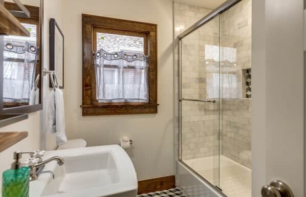 Un baño con cabina de ducha de vidrio, un lavabo blanco y una ventana con cortina.
