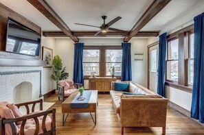 Un salón amplio con chimenea, piso de madera, ventilador de techo y ventanas grandes con cortinas azules.