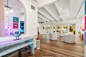 A spacious room with a wooden herringbone floor, white walls, and a large arched mirror. There are two white chairs, a small table, and a neon flamingo sign.