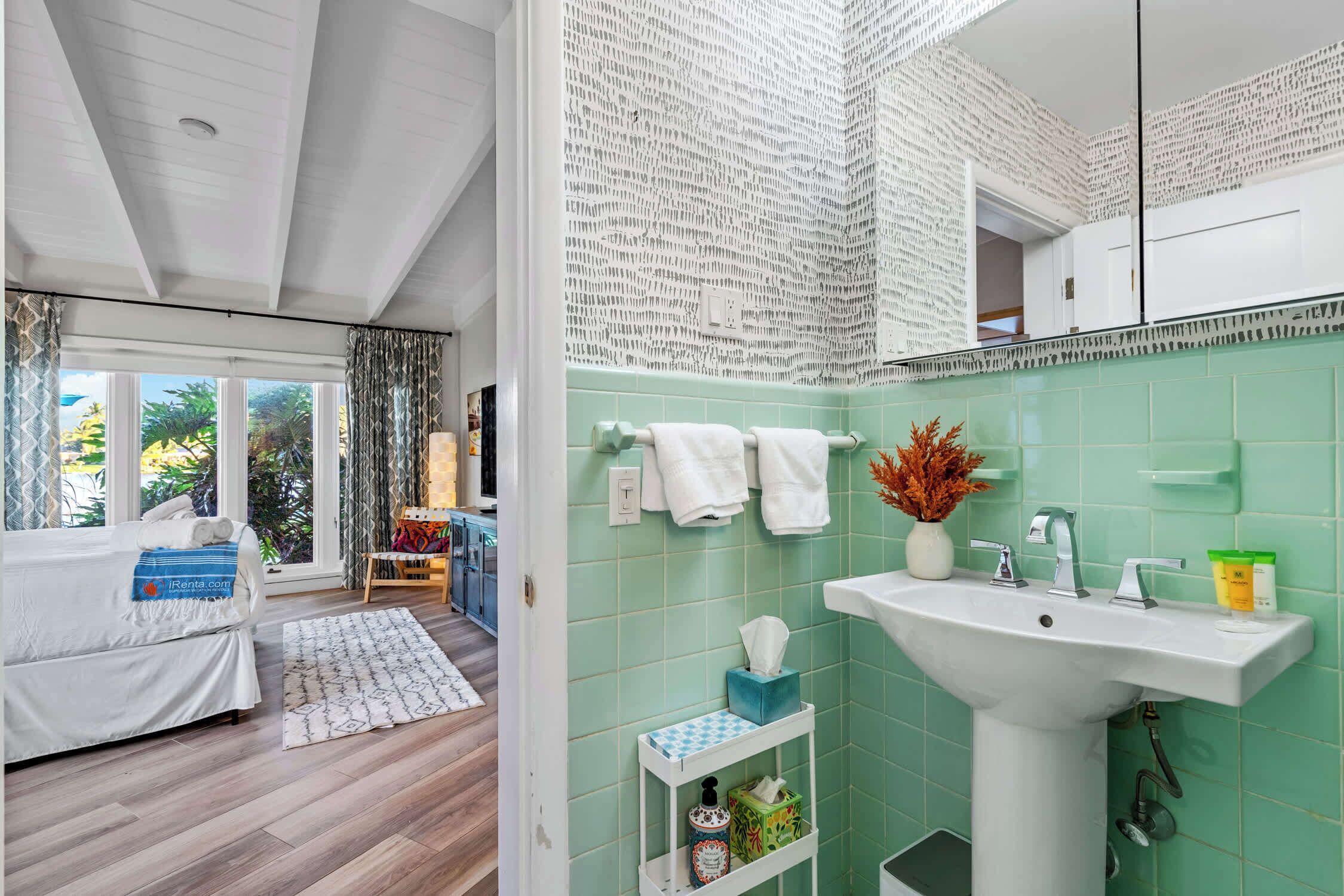 A modern hotel room with a large bed, a view of the outdoors, a bathroom with a green tiled wall, and a sink with toiletries.