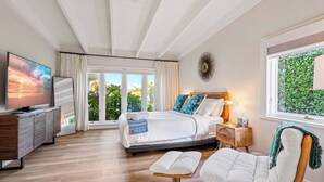 A modern bedroom with a bed, a TV, a wooden bench, and a window with a view of greenery. - The Clarke Collection-Villa Flamingo - Waterfront (Lake Clarke Shores)
