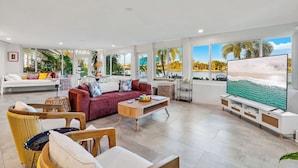 A spacious living room with a large TV, comfortable seating, and large windows with a view of palm trees. - The Clarke Collection-Villa Flamingo - Waterfront (Lake Clarke Shores)