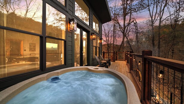 A hot tub on a wooden deck with large windows overlooking a forested area.