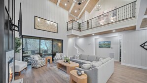 Un vaste espace de vie comprenant un canapé d’angle gris, des fauteuils à motifs et une table basse en bois. Une cheminée est visible sur la gauche, et une grande fenêtre offre une vue sur les arbres à l’extérieur.