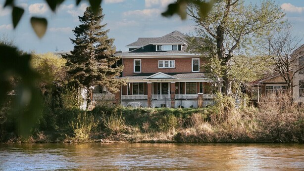 Ein zweistöckiges Haus mit Veranda, ein Fluss im Vordergrund und Bäume im Hintergrund.