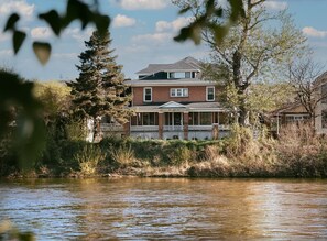 A two-story house with a porch, a river in the foreground, and trees in the background. - Apt. #5- Goldsmith's Riverfront Inn- Bitterroot Apartment (Missoula)