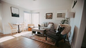 En moderne stue med trægulv, et sofabord, en sofa og en stol. Der er et vindue med persienner, en vægmonteret aircondition og et indrammet billede på væggen.