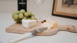 A wooden cutting board with a wedge of white cheese, a piece of soft cheese, and a bunch of green grapes. A knife rests on the board. - Apt. #6 · Goldsmith's River Front Inn - Rock Creek Apartment (Missoula)