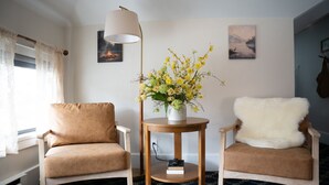 A room with two beige armchairs, a round wooden table with a vase of yellow flowers, a floor lamp, and two wall-mounted paintings. - Apt. #6 · Goldsmith's River Front Inn - Rock Creek Apartment (Missoula)