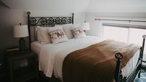 A neatly made bed with white linens and decorative pillows, a dark metal headboard, a bedside lamp, and a window with a view. - Apt. #6 · Goldsmith's River Front Inn - Rock Creek Apartment (Missoula)