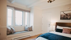 A bedroom with a large bed, a window seat with cushions, a door, and a painting on the wall.